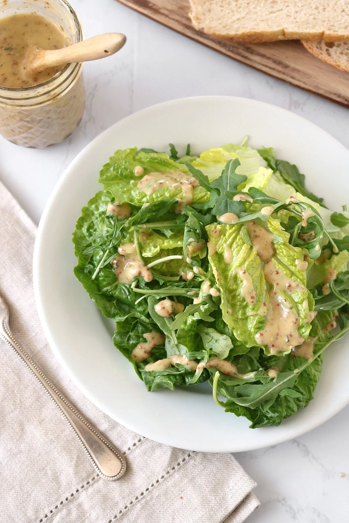 A white plate with fresh green lettuce and arugula salad drizzled with a creamy vinaigrette dressing. A jar of dressing, a folded napkin with a fork, and sliced bread are nearby on a marble surface.