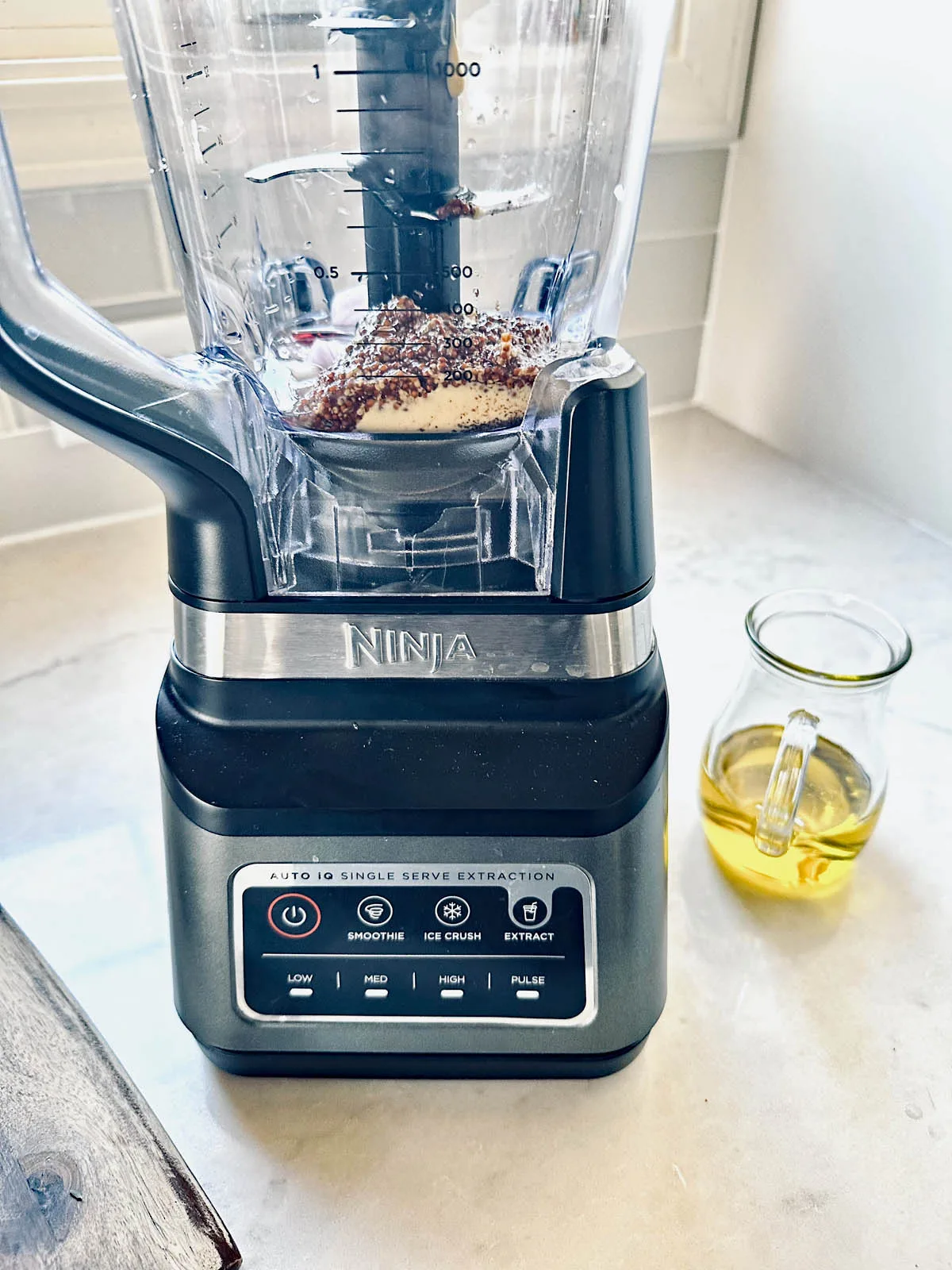 A Ninja blender on a countertop contains various spices and ingredients. Beside it sits a small glass pitcher filled with yellow oil. The blenders control panel is visible, showing several blending options.