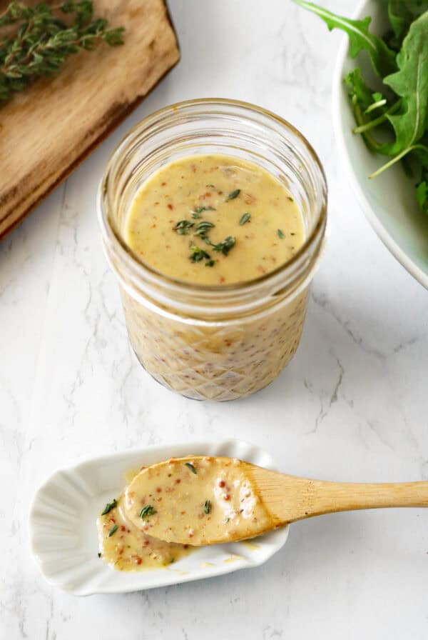 A small glass jar filled with creamy mustard vinaigrette sits on a white marble surface. Beside it, a wooden spoon rests on a white dish, holding a sample of the sauce. Fresh herbs and greens are nearby.