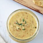 A glass jar filled with creamy mustard sauce garnished with fresh thyme, placed on a white marble surface near a wooden board and a spoon with sauce.