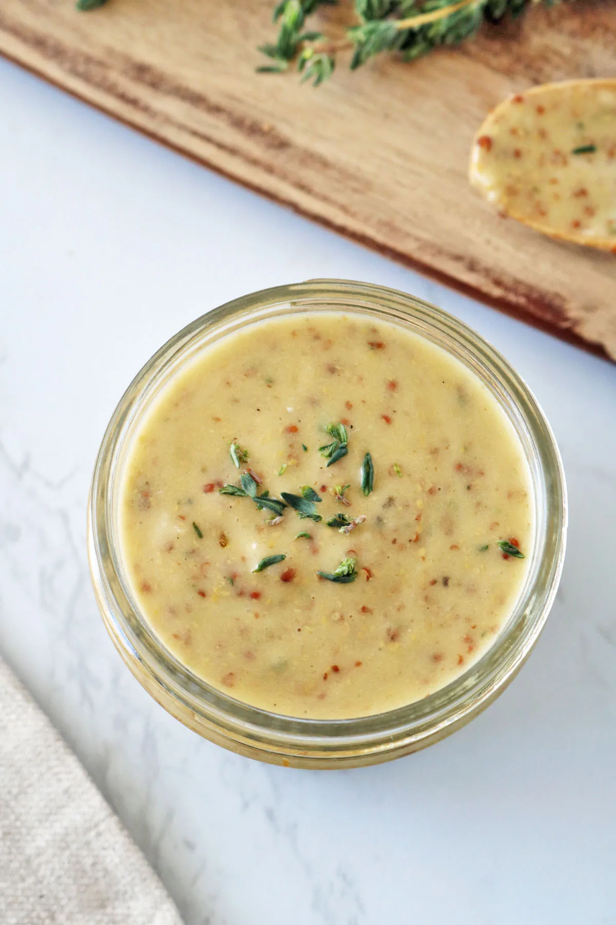 A glass jar filled with creamy mustard sauce garnished with fresh thyme, placed on a white marble surface near a wooden board and a spoon with sauce.