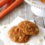 Two carrot cookies with white chocolate chips rest on a white napkin, with fresh carrots and more cookies displayed on a cake stand in the background on a rustic wooden surface.