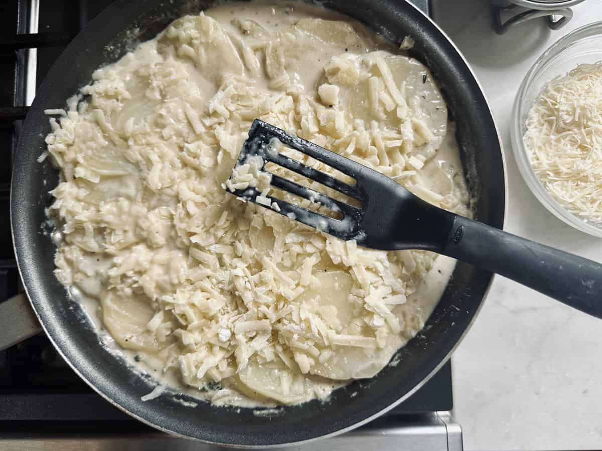 A skillet on a stove contains sliced potatoes in a creamy sauce, topped with shredded cheese. A black spatula rests in the pan, and a bowl of additional shredded cheese sits nearby on the counter.