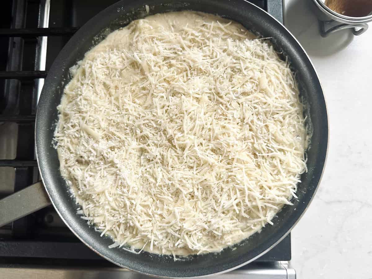 A frying pan on a stove filled with uncooked dough covered in a generous layer of shredded cheese, ready to be cooked. A small container of brown powder is visible on the countertop nearby.