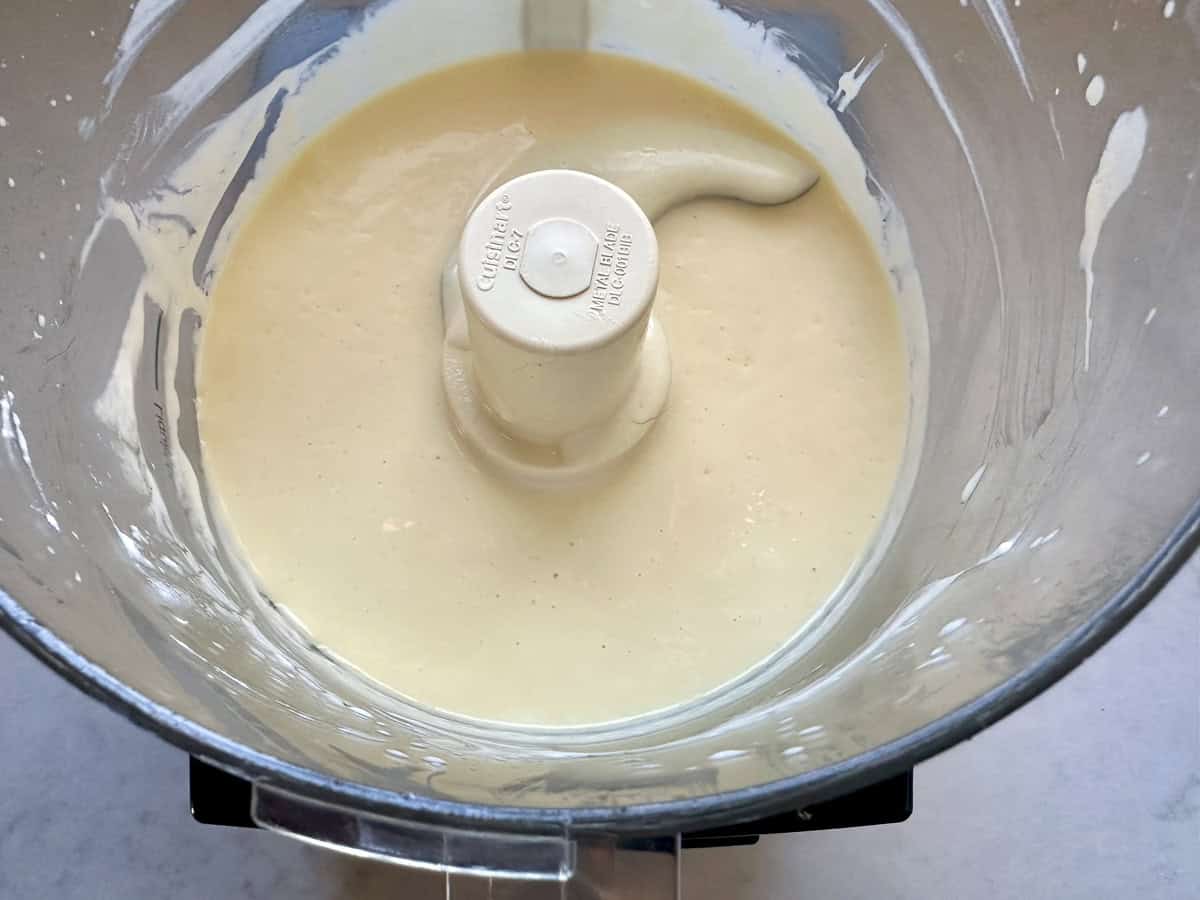A food processor bowl filled with a smooth, creamy batter, with some batter coating the sides and the central blade attachment visible in the middle.