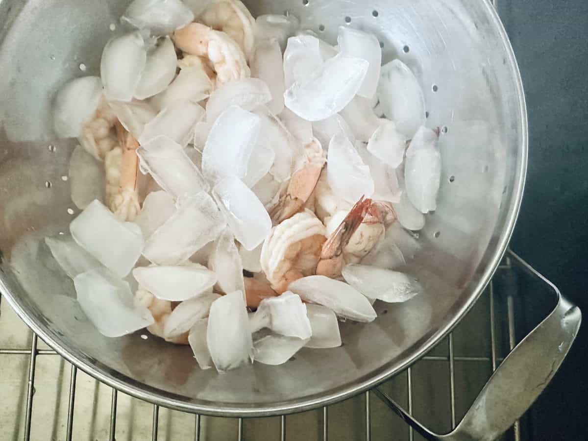 A metal colander filled with cooked shrimp and ice cubes sits on a wire rack over a sink, chilling the shrimp.