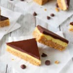 Two triangular Chocolate-Glazed Crispy Peanut Butter Pudding Bars with layers of chocolate, creamy yellow filling, and a crumbly base rest on white parchment paper. A few chocolate chips are scattered nearby on a white wooden surface.