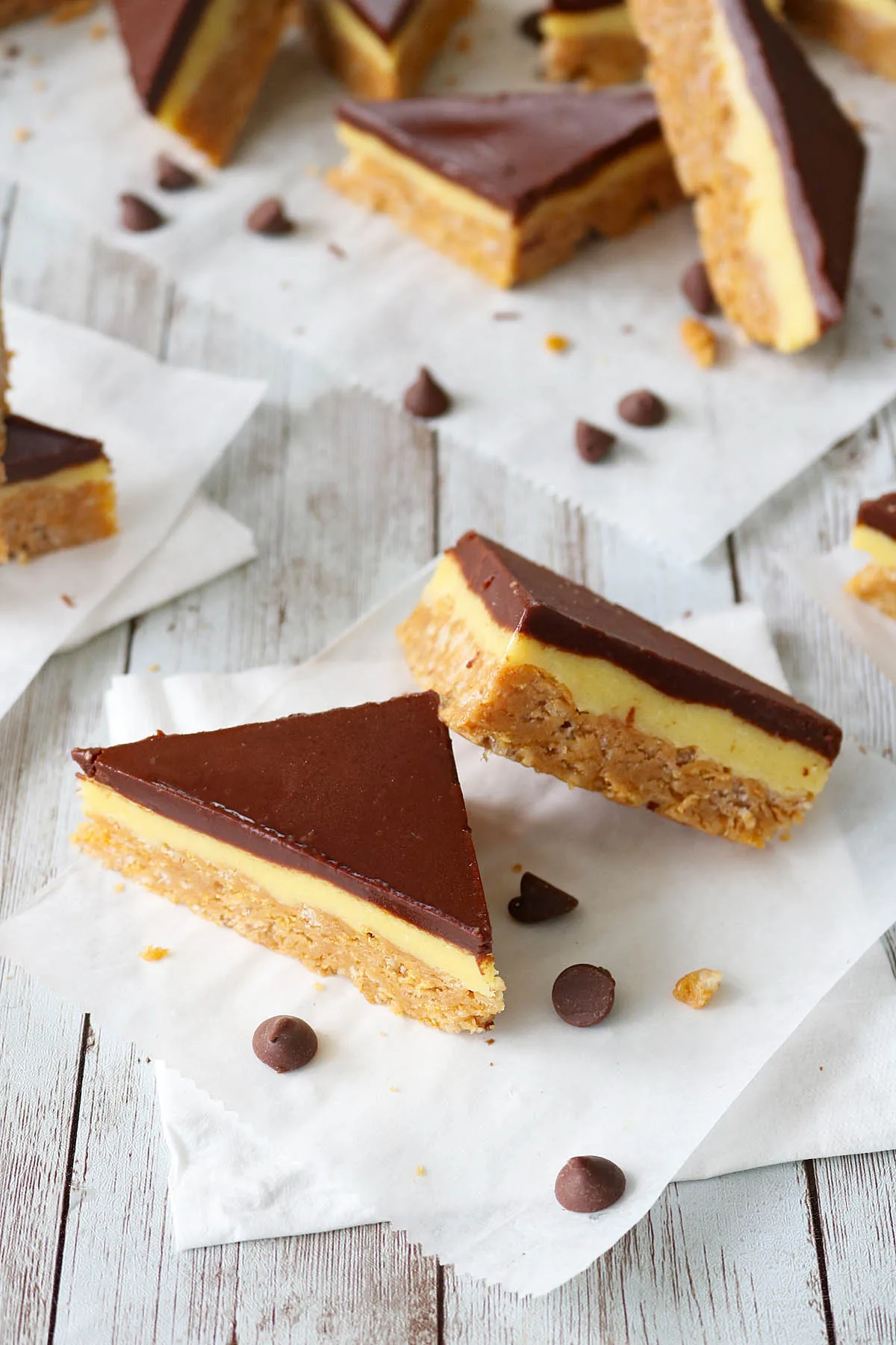 Two triangular Chocolate-Glazed Crispy Peanut Butter Pudding Bars with layers of chocolate, creamy yellow filling, and a crumbly base rest on white parchment paper. A few chocolate chips are scattered nearby on a white wooden surface.