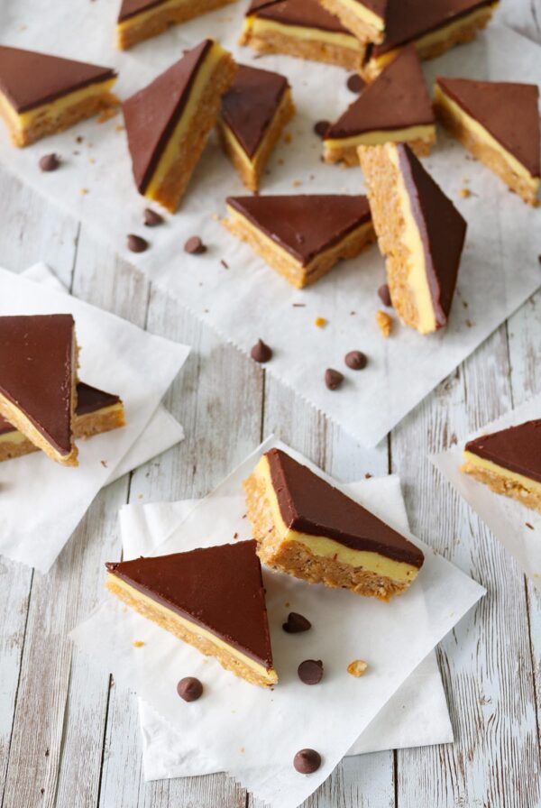 Several Chocolate-Glazed Crispy Peanut Butter Pudding Bars are arranged on parchment paper over a rustic white wooden surface, with a few chocolate chips scattered around.