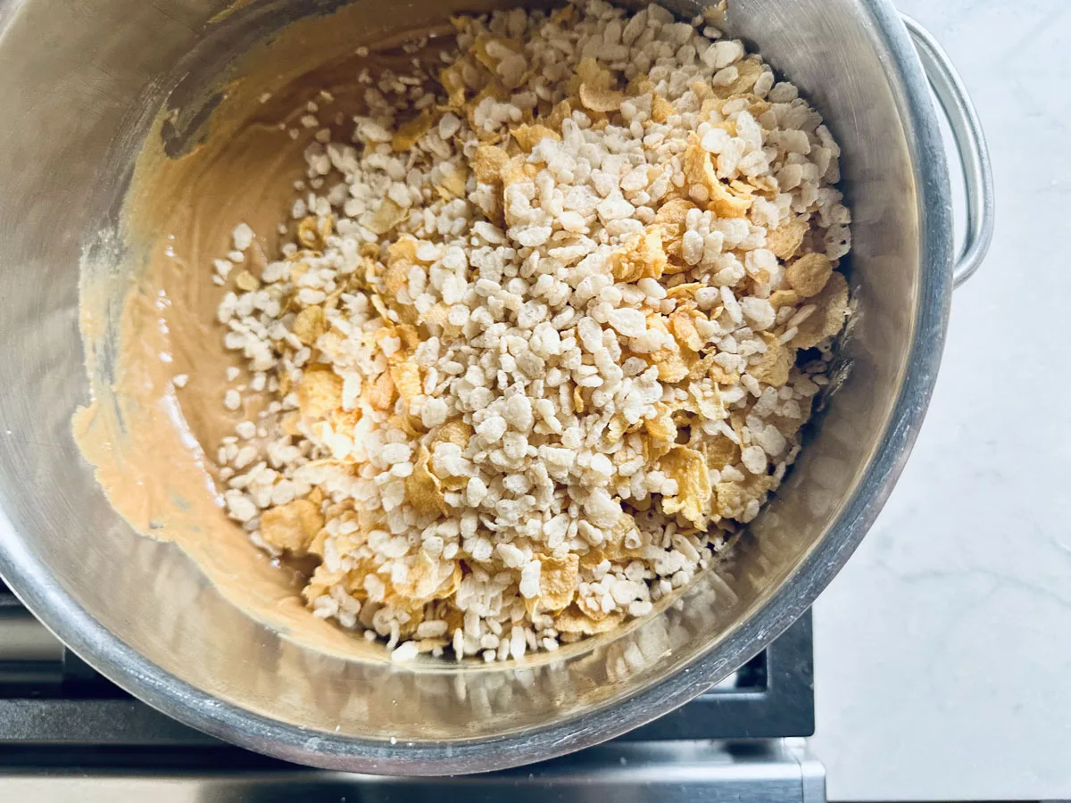 A stainless steel mixing bowl filled with a mixture of creamy batter, puffed rice cereal, and cornflakes sits on a stovetop.