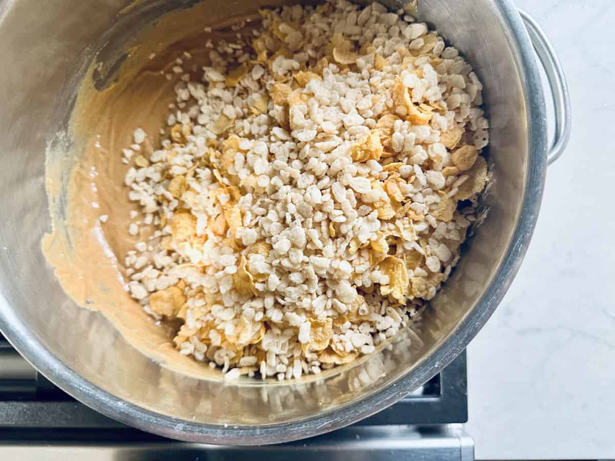 A stainless steel mixing bowl filled with a mixture of creamy batter, puffed rice cereal, and cornflakes sits on a stovetop.