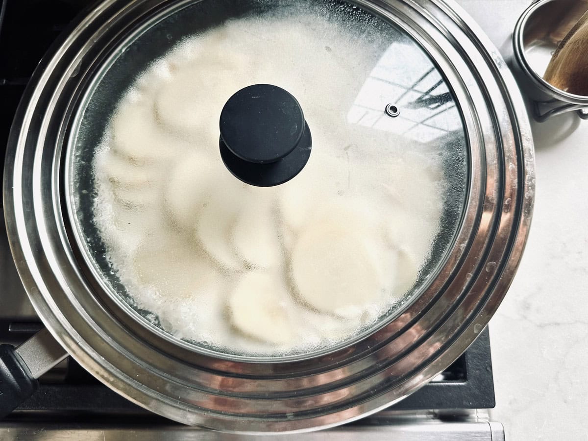 A pot with a glass lid steaming on a stovetop, showing round dumplings or dough pieces inside being cooked; a metal cup with a brush is nearby on a light-colored countertop.