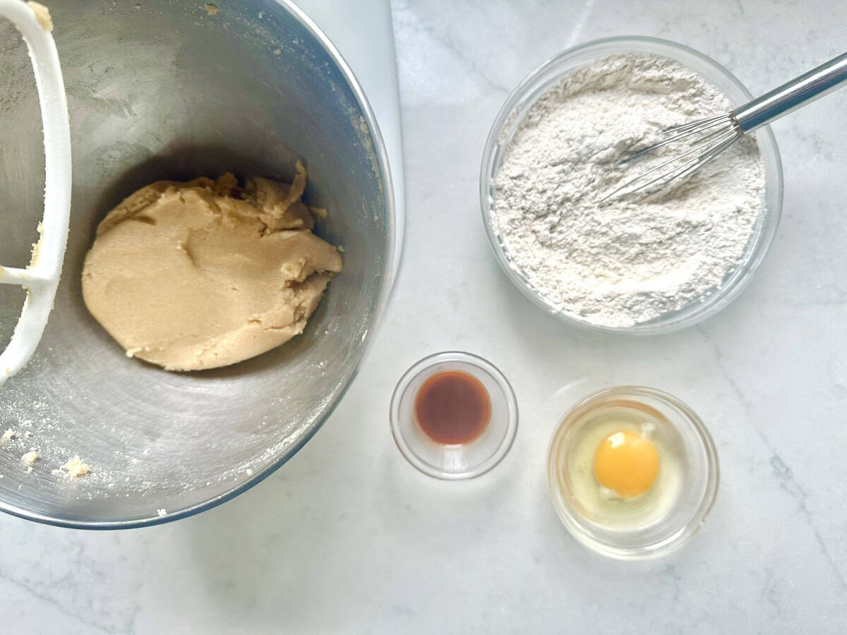 A mixing bowl with cookie dough, a bowl of flour with a whisk, a small bowl of vanilla extract, and a small bowl with a cracked egg are arranged on a white countertop.