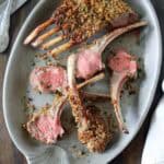 An oval platter with Crumb-Crusted Rack of Lamb Persillade, each slice revealing pink centers beneath a golden herb and breadcrumb crust. A fork and knife rest nearby on the dark wooden table.