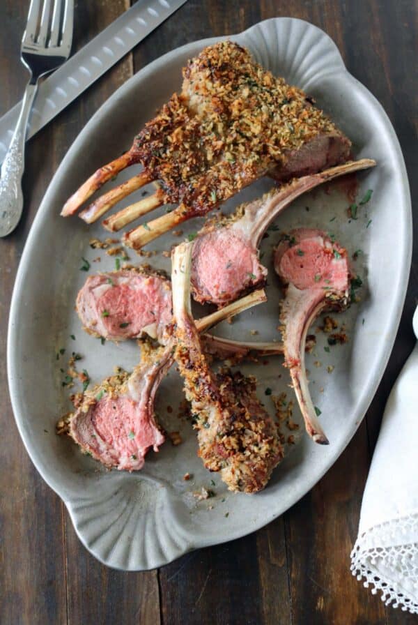 An oval platter with Crumb-Crusted Rack of Lamb Persillade, each slice revealing pink centers beneath a golden herb and breadcrumb crust. A fork and knife rest nearby on the dark wooden table.