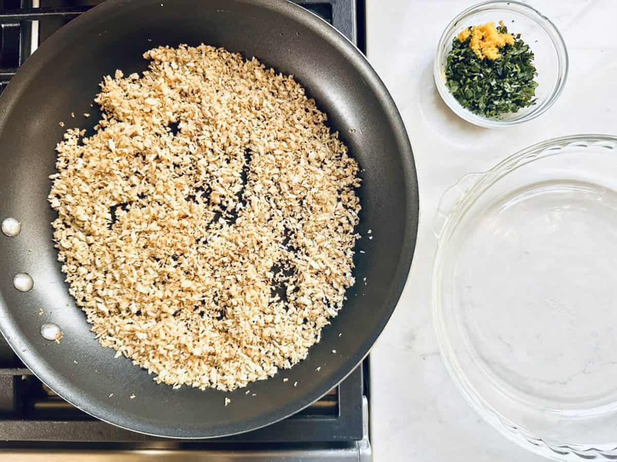 A skillet with toasted breadcrumbs for Crumb-Crusted Rack of Lamb Persillade sits on a stovetop, next to a small bowl of chopped herbs and grated orange peel, with an empty glass dish on a white countertop.
