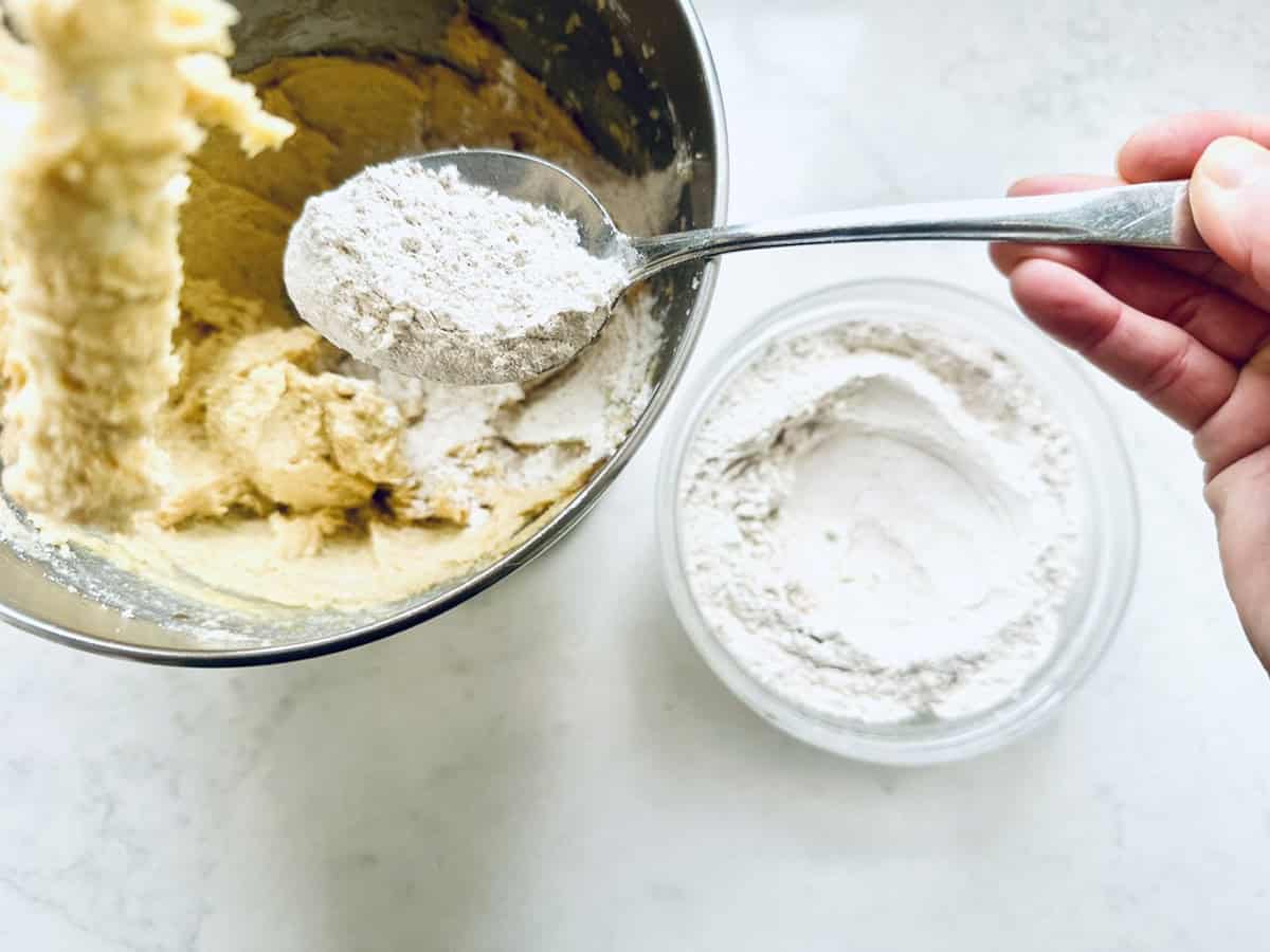 A hand holds a spoon of flour above a mixing bowl with yellow batter. A separate bowl of flour sits nearby on a white countertop.