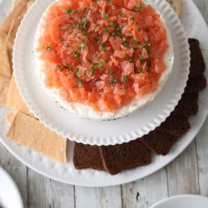 A plate with a round Gravlax Torta&mdash;smoked salmon and cream cheese dip topped with chives&mdash;surrounded by assorted crackers and bread slices on a rustic wooden table.