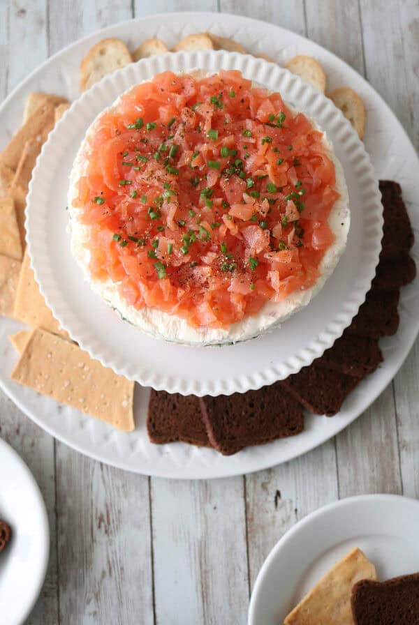 A plate with a round Gravlax Torta—smoked salmon and cream cheese dip topped with chives—surrounded by assorted crackers and bread slices on a rustic wooden table.