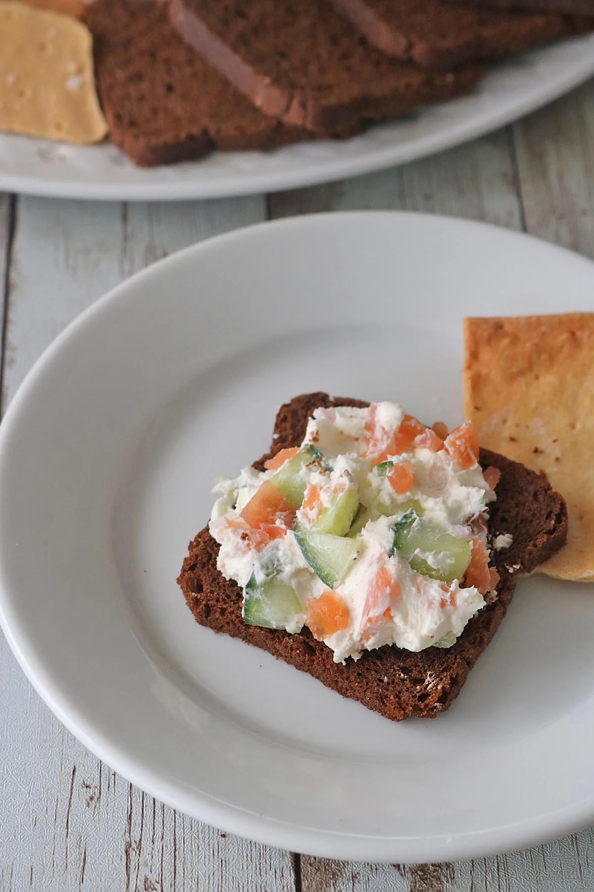 A slice of dark bread topped with a creamy Gravlax Torta spread containing chopped cucumbers and orange-colored vegetables, served on a white plate next to a cracker. More bread slices are visible in the background.