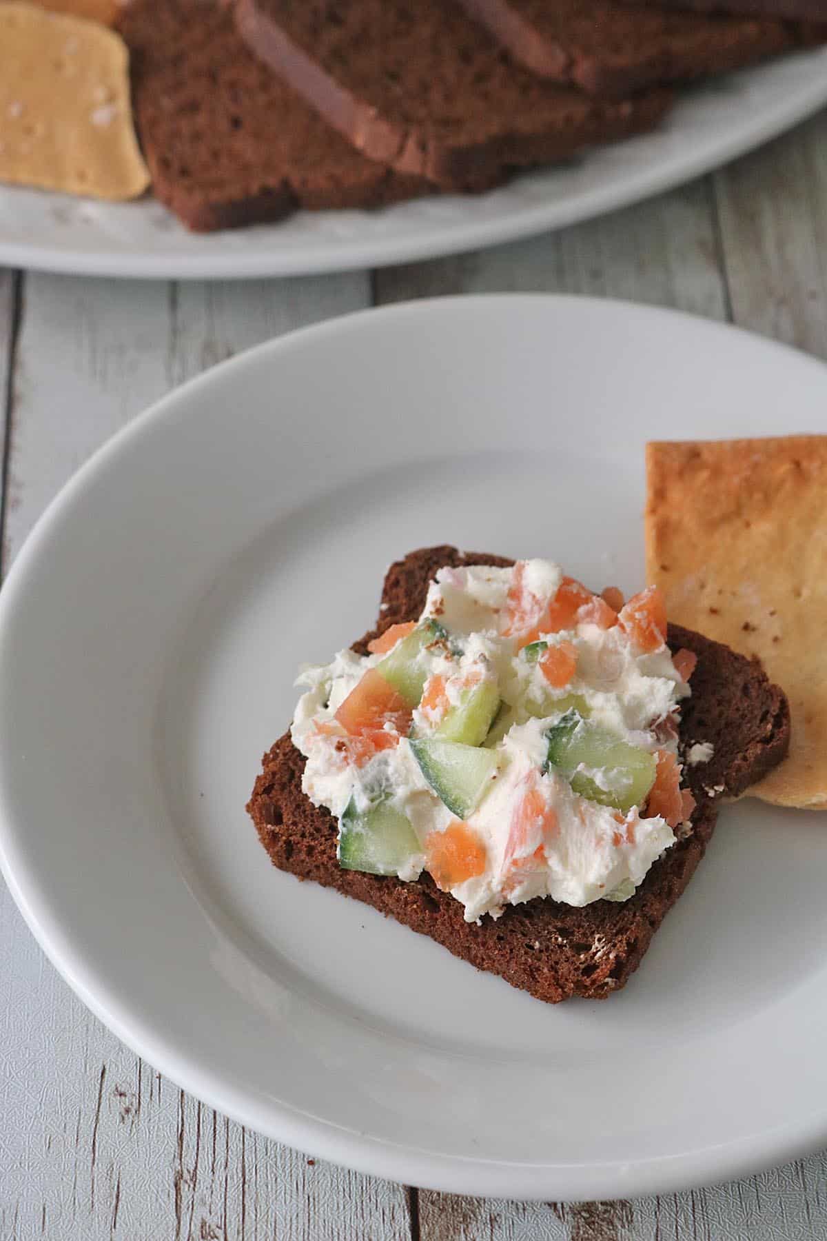 A slice of dark bread topped with a creamy Gravlax Torta spread containing chopped cucumbers and orange-colored vegetables, served on a white plate next to a cracker. More bread slices are visible in the background.