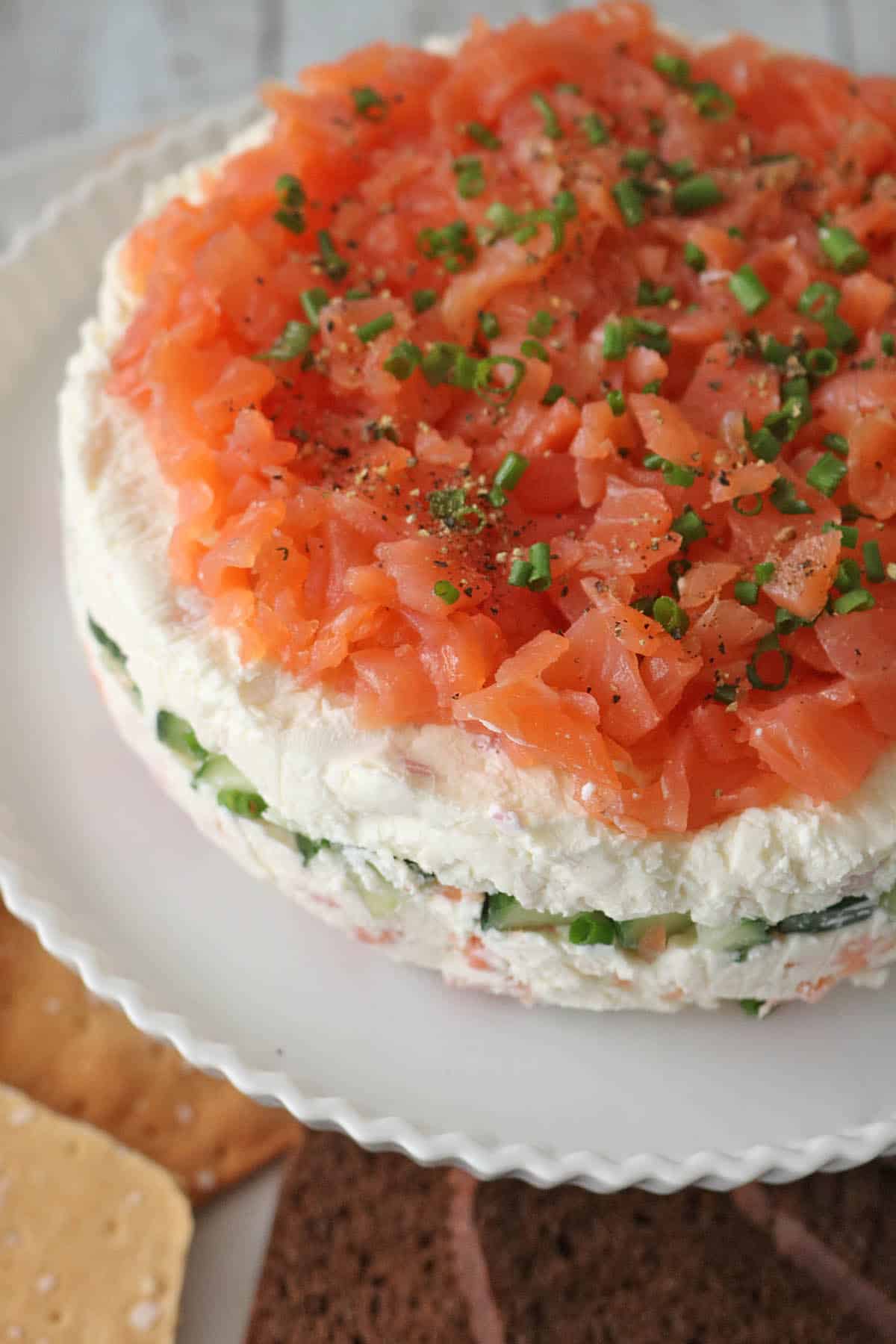 A round layered Gravlax Torta appetizer topped with diced smoked salmon and chopped chives, featuring visible layers of cream cheese and cucumber, served on a white plate with assorted crackers on the side.
