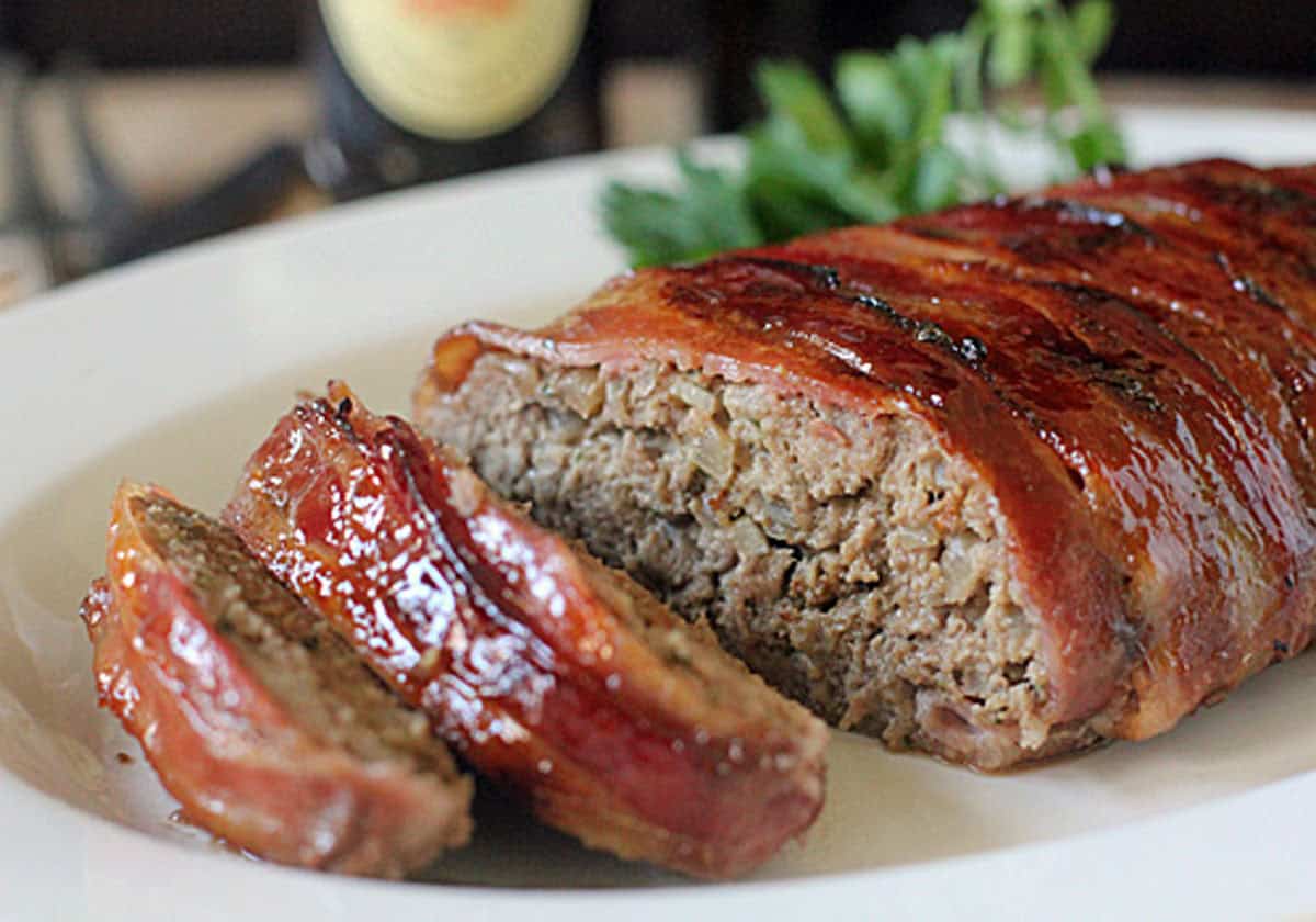 A bacon-wrapped meatloaf is sliced and served on a white plate, with fresh herbs in the background. The meatloaf has a glossy, caramelized glaze on top.