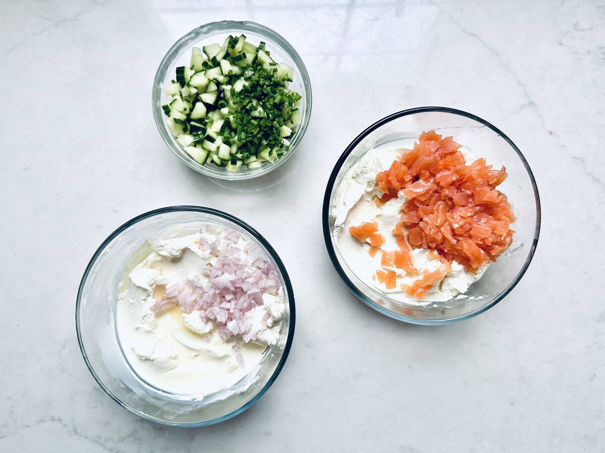Three glass bowls on a white surface: one with chopped cucumber and chives, one with cream cheese and chopped shallots, and one with cream cheese and smoked salmon—perfect components for a delicious Gravlax Torta.