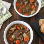 Two white bowls filled with lamb stew containing chunks of lamb, carrots, and potatoes, garnished with herbs, are on a wooden table next to a sliced baguette and napkins with a spoon.