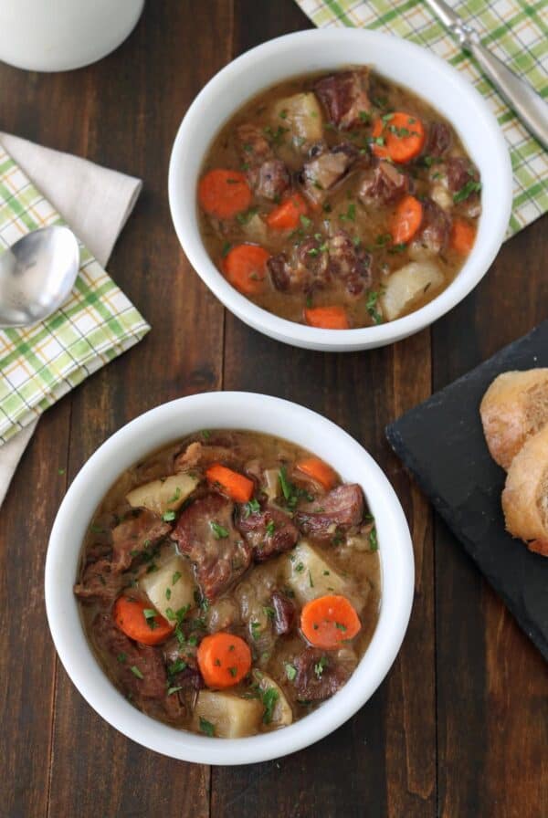 Two white bowls filled with lamb stew containing chunks of lamb, carrots, and potatoes, garnished with herbs, are on a wooden table next to a sliced baguette and napkins with a spoon.