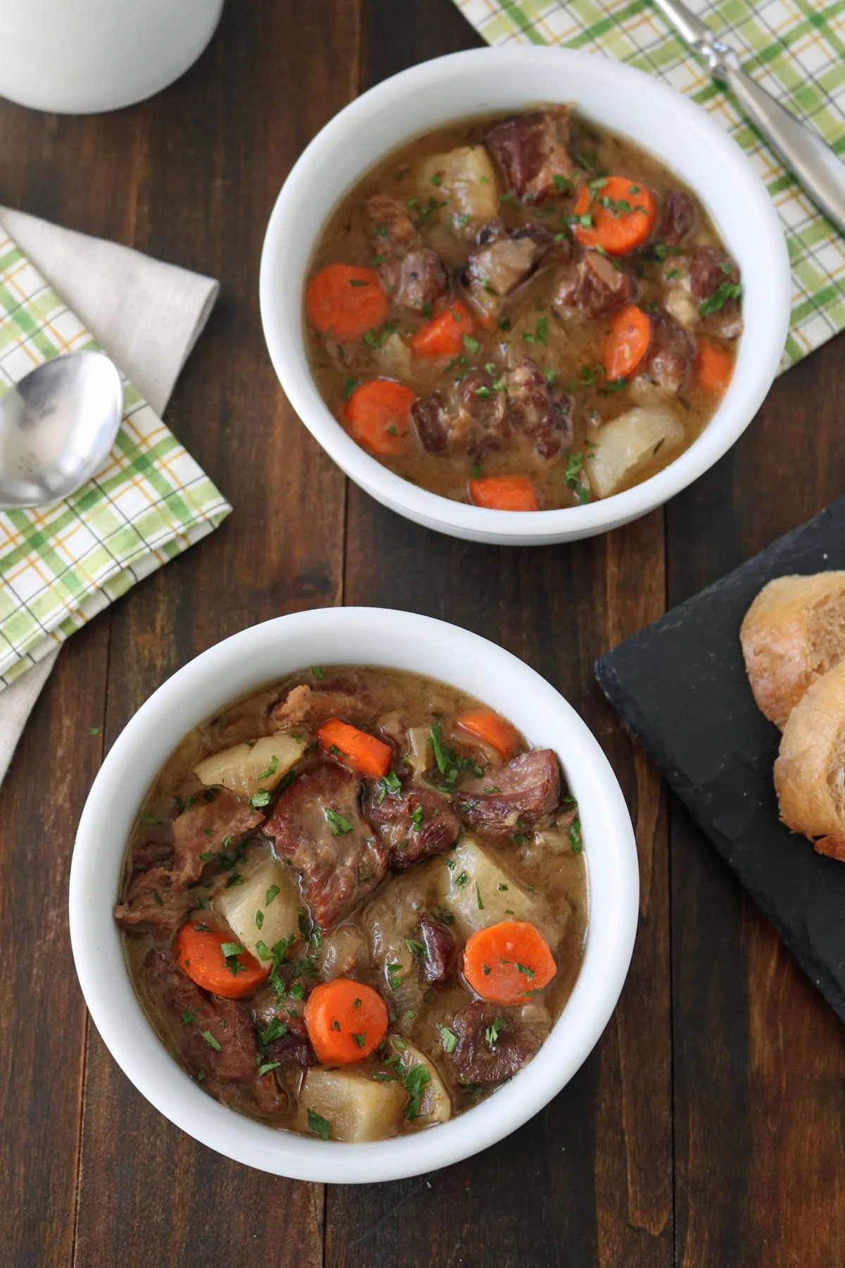 Two white bowls filled with lamb stew containing chunks of lamb, carrots, and potatoes, garnished with herbs, are on a wooden table next to a sliced baguette and napkins with a spoon.