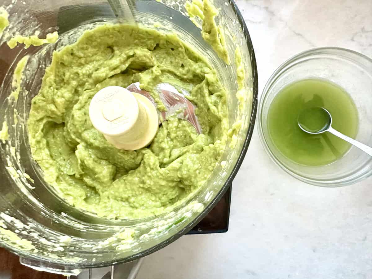 A food processor filled with creamy mashed avocado sits next to a glass bowl containing green liquid with a spoon in it, all on a light-colored countertop.
