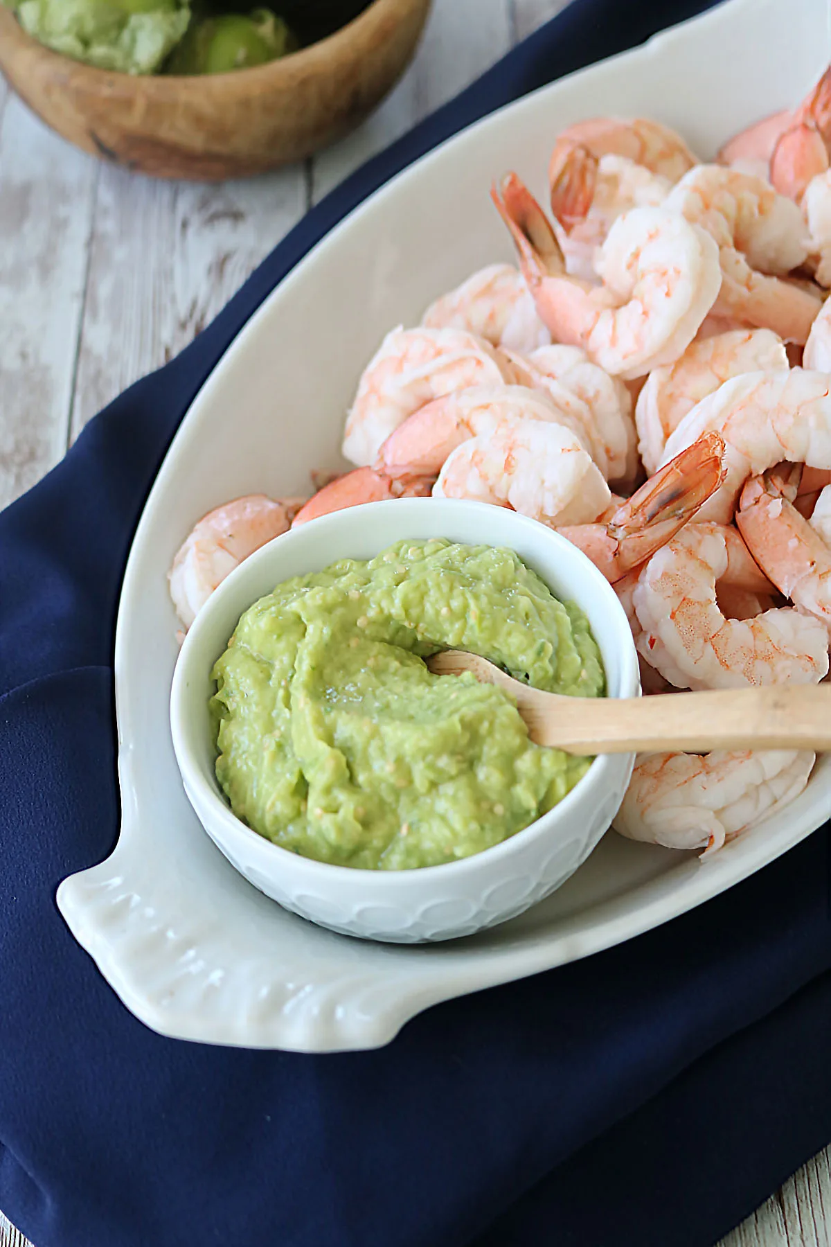 A white serving dish filled with cooked shrimp, next to a small bowl of guacamole with a wooden spoon, placed on a dark blue cloth.