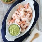 A white oval platter with cooked shrimp and a small bowl of guacamole sits on a navy cloth. Nearby are a wooden bowl with avocado, napkins, and a wooden spoon on a rustic wooden surface.