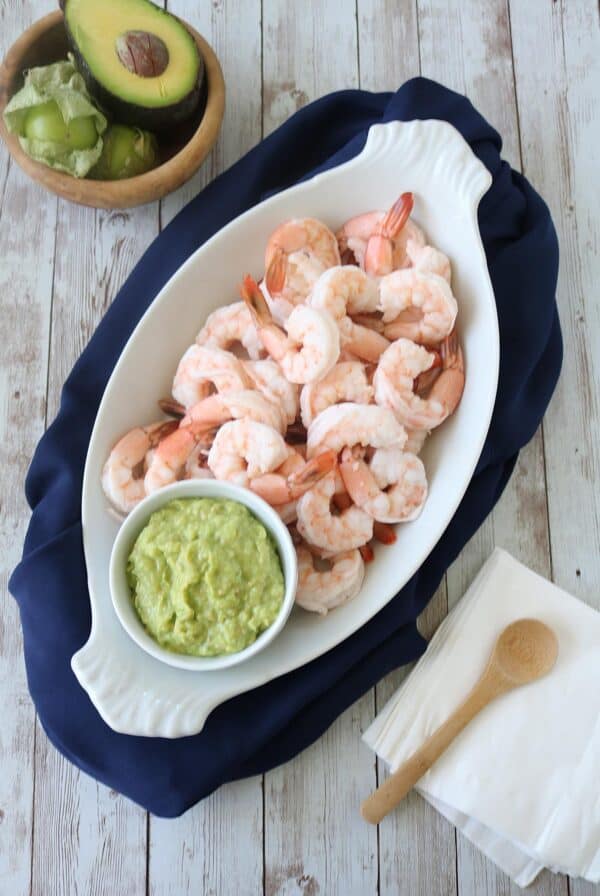 A white oval platter with cooked shrimp and a small bowl of guacamole sits on a navy cloth. Nearby are a wooden bowl with avocado, napkins, and a wooden spoon on a rustic wooden surface.