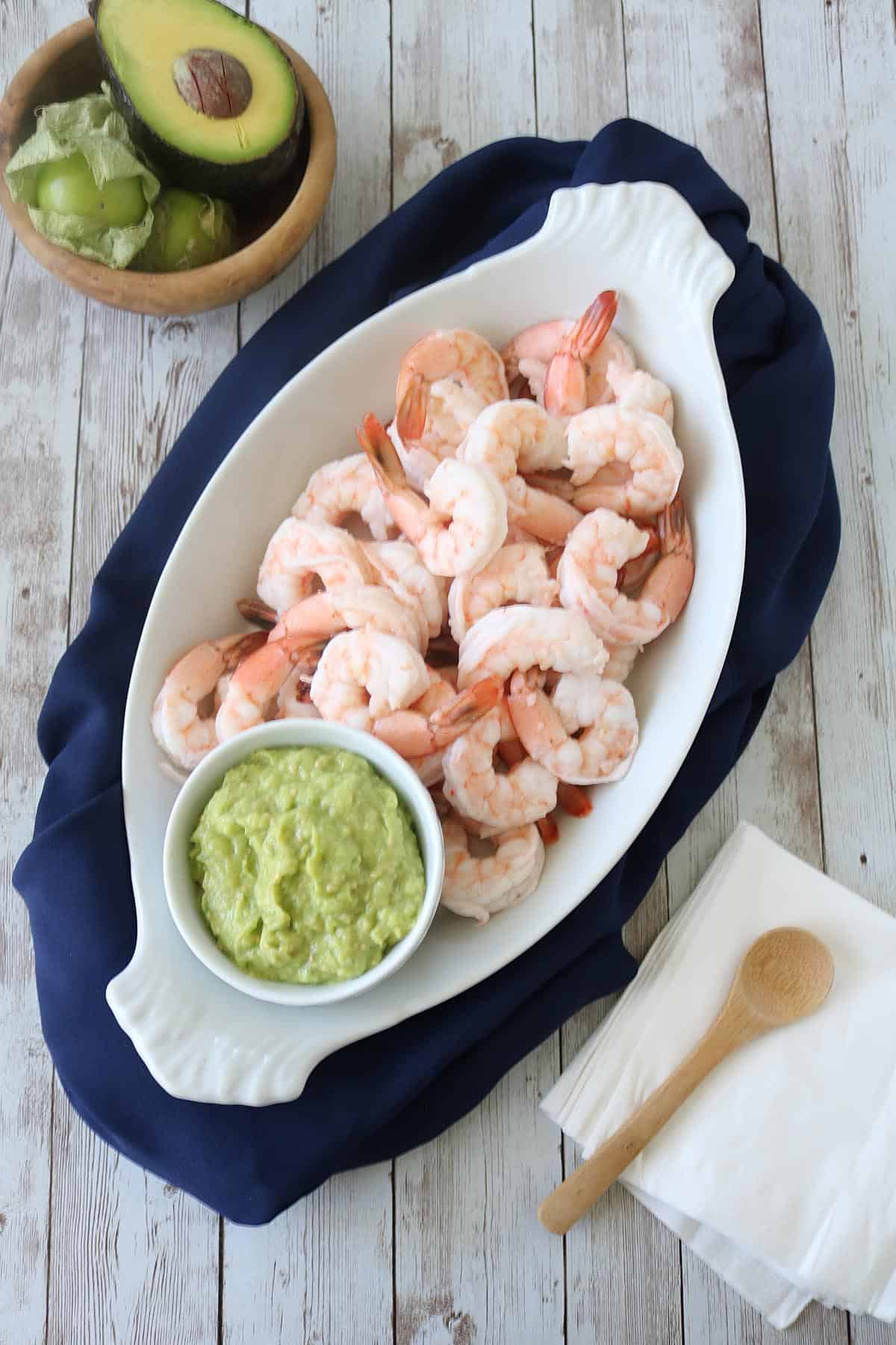 A white oval platter with cooked shrimp and a small bowl of guacamole sits on a navy cloth. Nearby are a wooden bowl with avocado, napkins, and a wooden spoon on a rustic wooden surface.