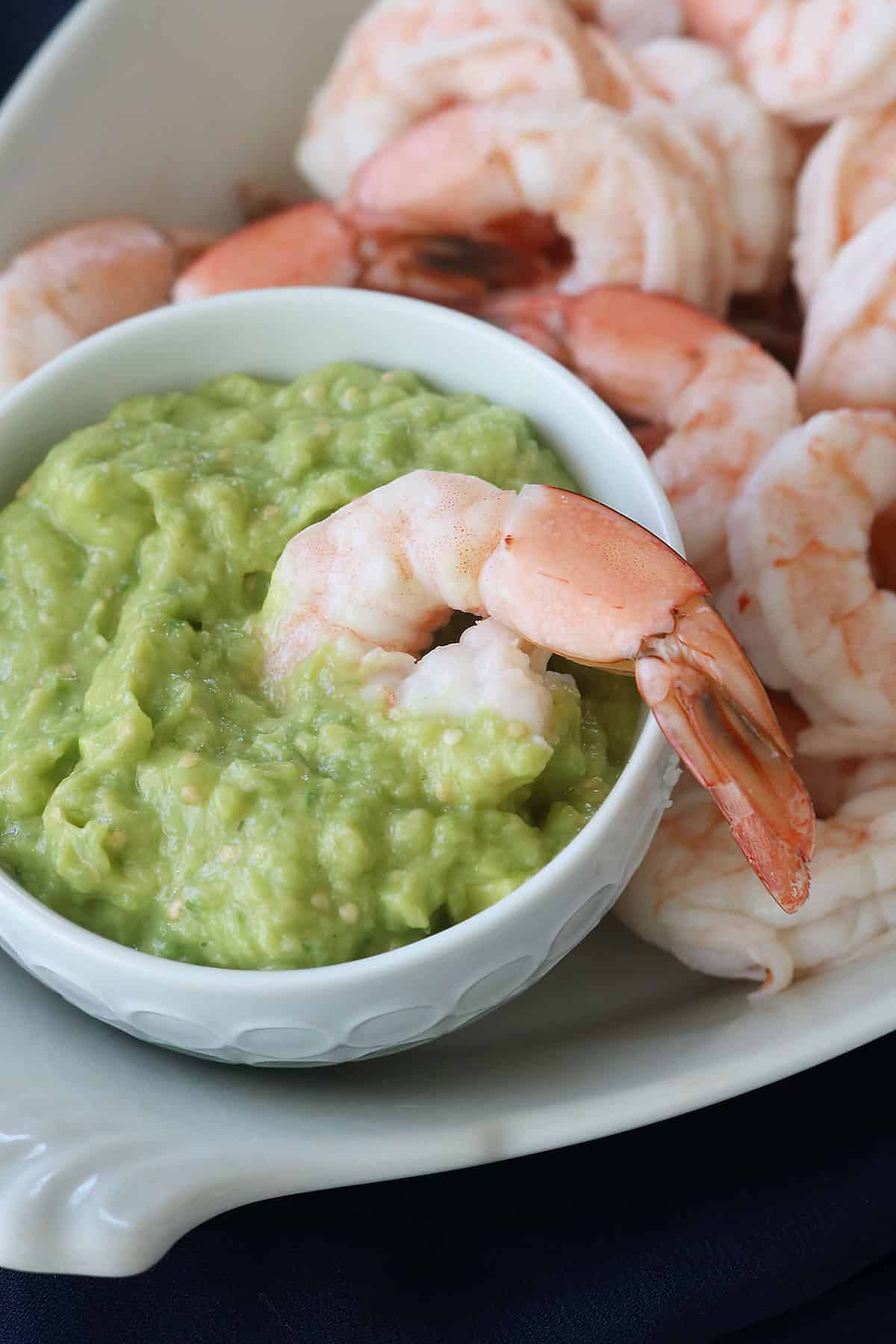 A bowl of guacamole with a cooked shrimp resting on its edge, surrounded by more shrimp on a white serving dish.