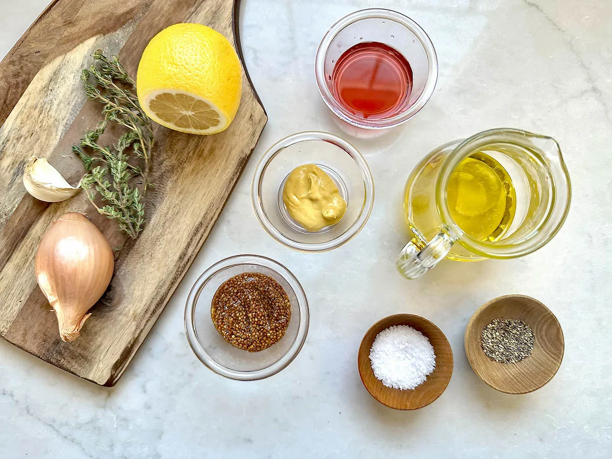 Ingredients for a vinaigrette arranged on a white surface: half a lemon, thyme, a garlic clove, a shallot, red wine vinegar, Dijon mustard, whole grain mustard, olive oil, salt, and black pepper.