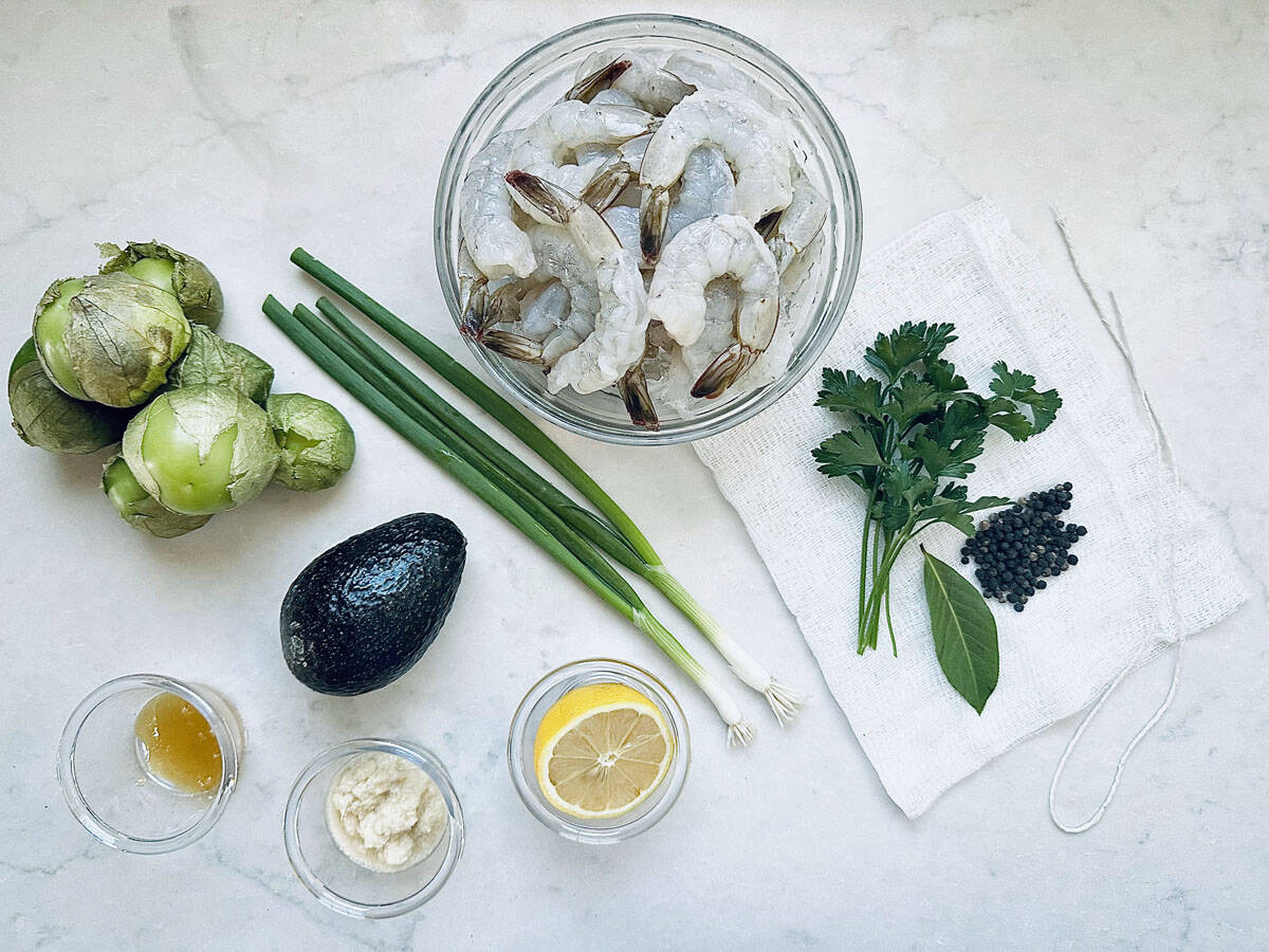 A bowl of raw shrimp, tomatillos, green onions, avocado, lemon slice, parsley, bay leaf, capers, horseradish, and a small dish of sauce arranged on a white surface with a cloth napkin.