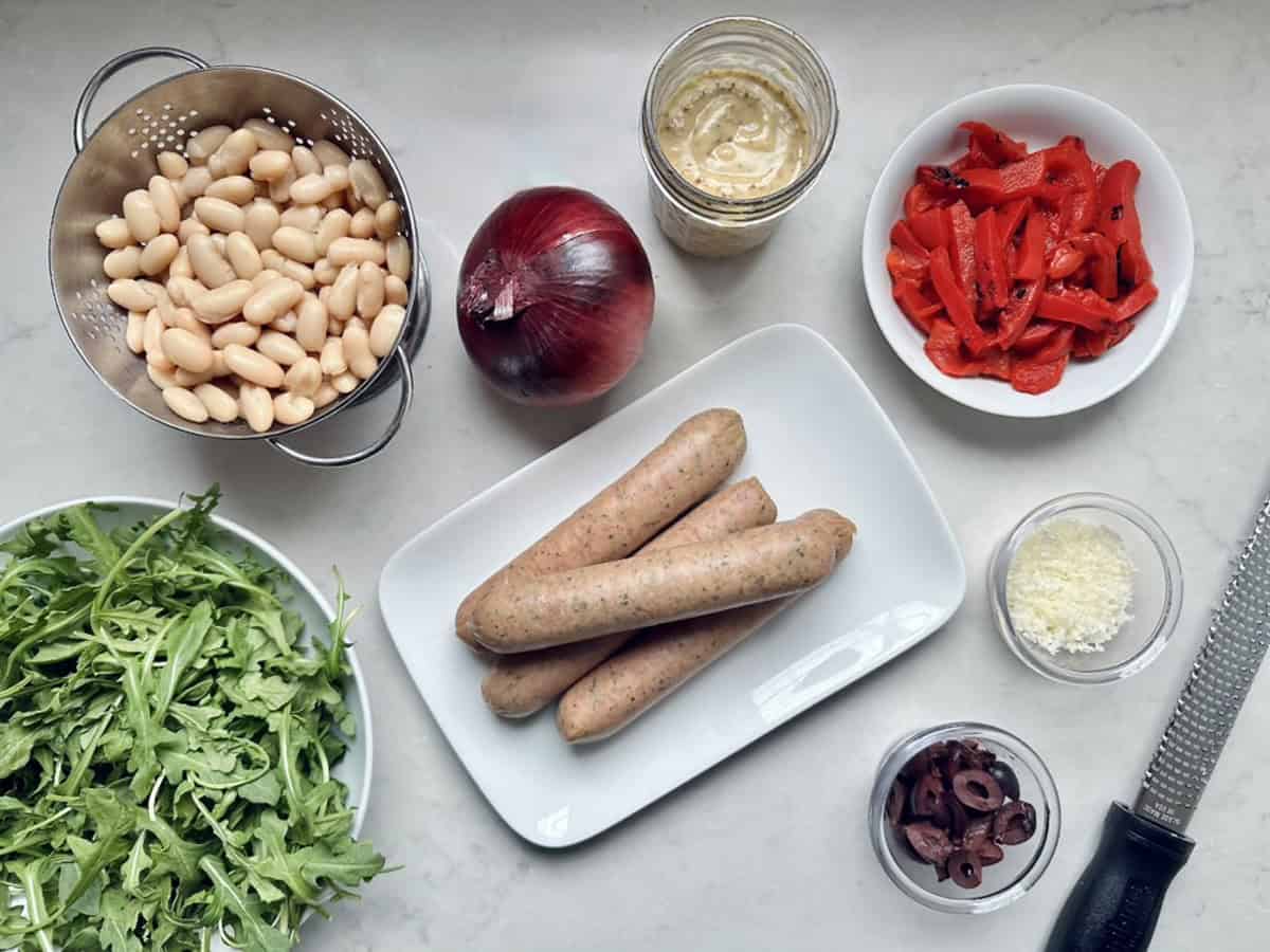 A top-down view of arugula salad ingredients on a countertop: cannellini beans, arugula, red onion, sausages on a plate, jar of mustard, sliced roasted red peppers, grated cheese, and sliced black olives—perfect for quick salad meals.