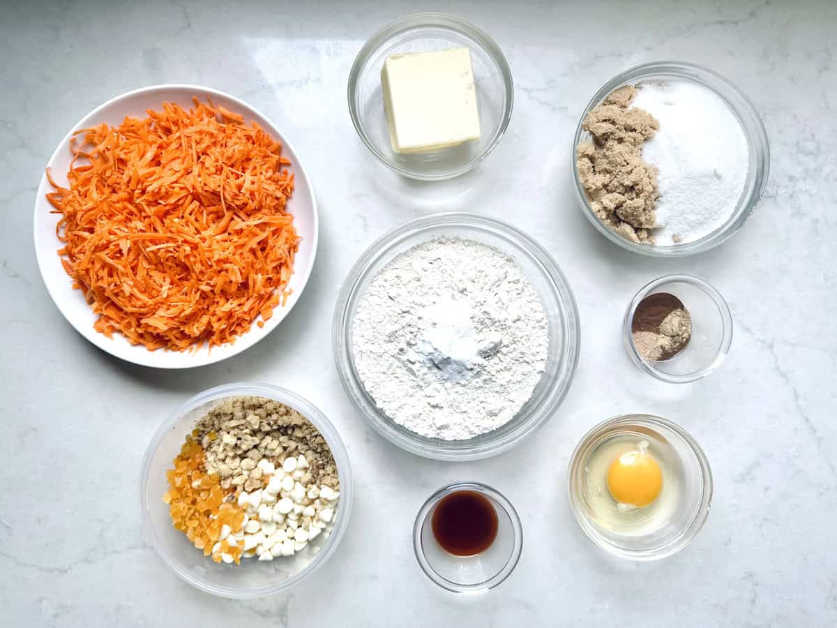 Various baking ingredients in bowls on a white surface, including shredded carrots, butter, flour with baking powder, brown and white sugars, cinnamon, nutmeg, walnuts, dried fruit, white chocolate chips, vanilla, and an egg.
