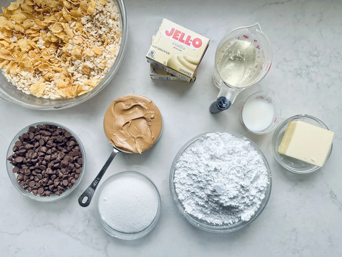 Various baking ingredients on a white surface, including a bowl of cornflakes, Rice Krispies, chocolate chips, a cup of peanut butter, sugar, powdered sugar, vanilla Jello pudding mix, butter, milk, and a measuring cup with liquid.