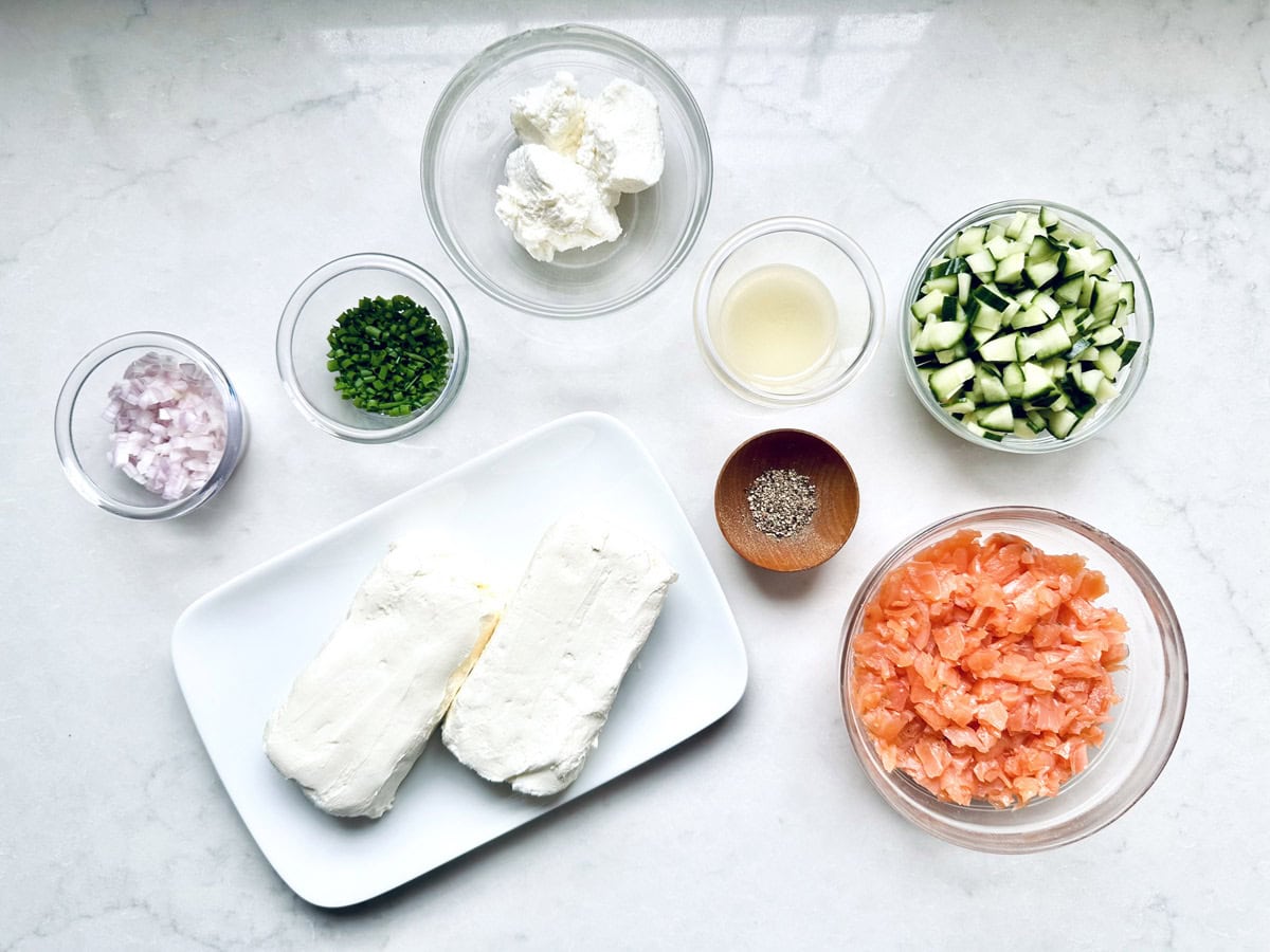 Ingredients on a white surface: two blocks of cream cheese, small bowls of diced smoked salmon, chopped cucumber, chives, diced shallots, lemon juice, and ground pepper—perfect for creating a flavorful Gravlax Torta.