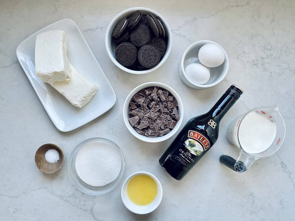 A top-down view of ingredients on a countertop: cream cheese, chocolate sandwich cookies, eggs, dark chocolate chunks, Baileys Irish Cream, milk, melted butter, sugar, and a small bowl of salt.