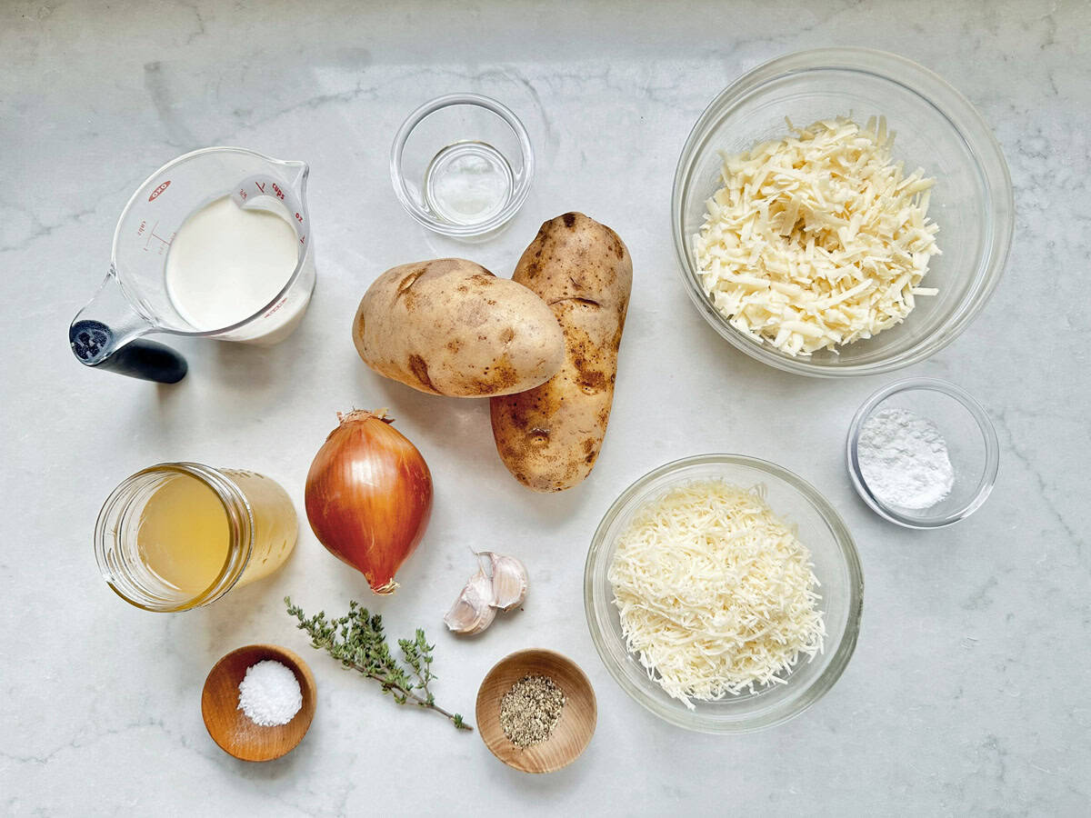 Overhead view of ingredients on a white surface: two russet potatoes, shredded cheese in two bowls, milk in a measuring cup, an onion, two garlic cloves, fresh thyme, chicken broth in a jar, flour, salt, and pepper in small bowls.