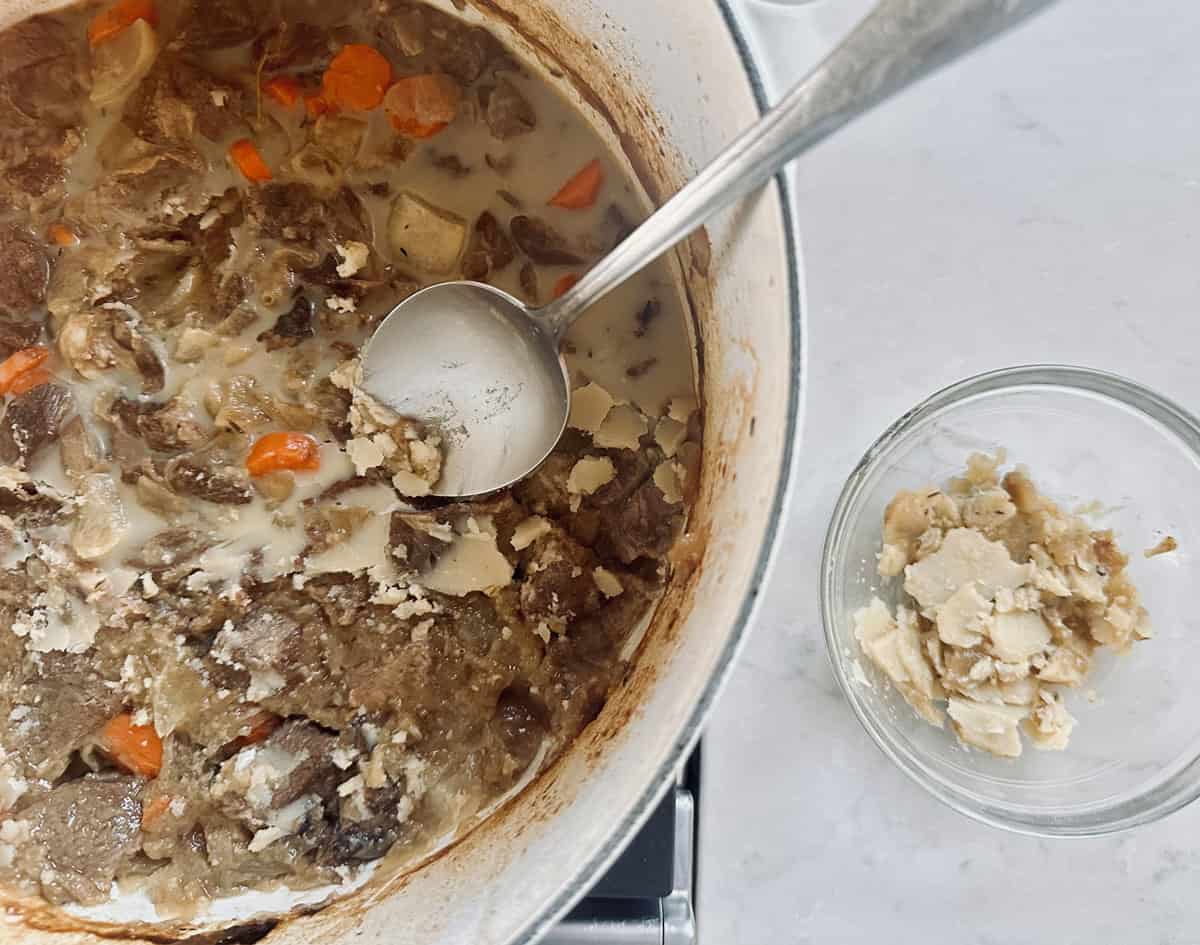 A pot of beef stew with carrots and potatoes, topped with a layer of solidified fat. A metal ladle is scooping out fat, which is collected in a small glass bowl on the side.