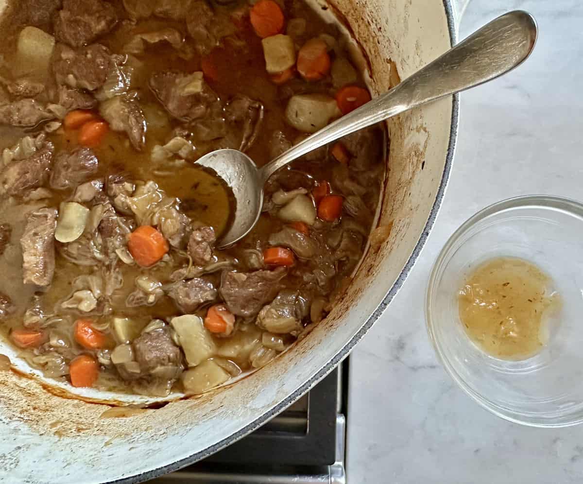 A pot of beef stew with chunks of meat, carrots, and potatoes sits on a stove next to a bowl containing a small amount of broth. A spoon rests inside the pot.