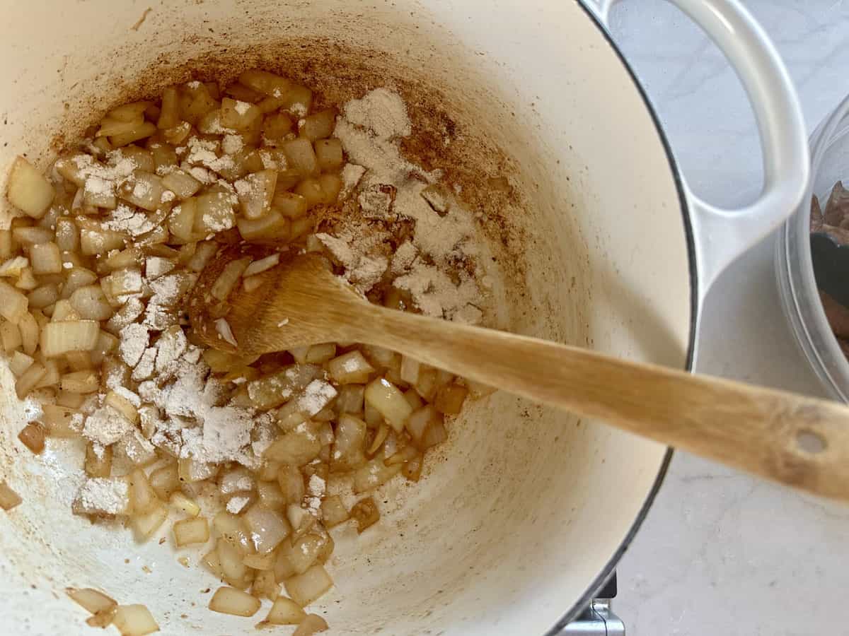 Chopped onions cooking in a large white pot, partially browned, with flour sprinkled on top and a wooden spoon stirring them. The pot sits on a white surface next to a clear bowl.