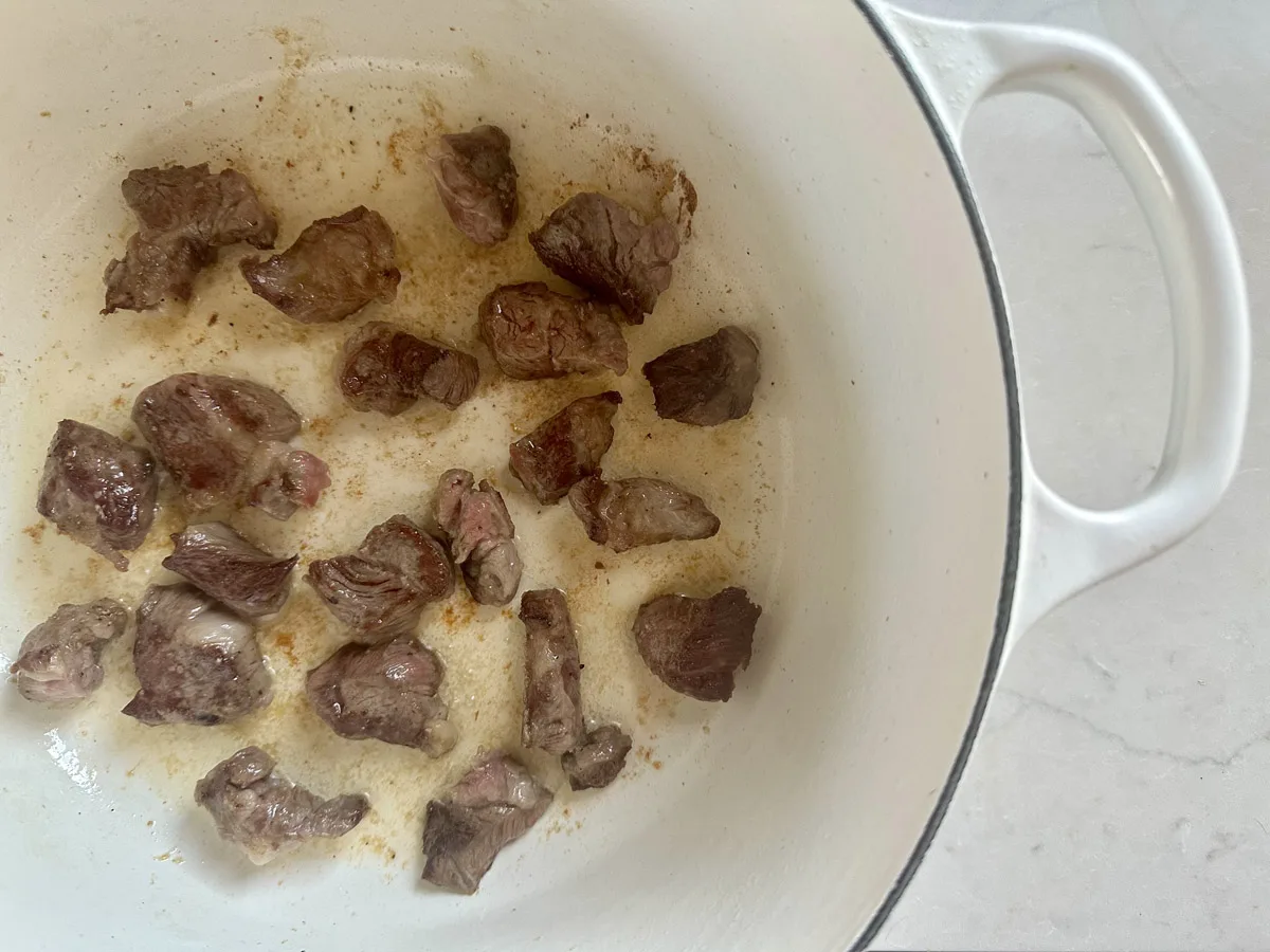 Chunks of beef are browning in a white enameled pot on a light-colored countertop. The meat is seared, and some oil or fat is visible around the pieces in the pot.
