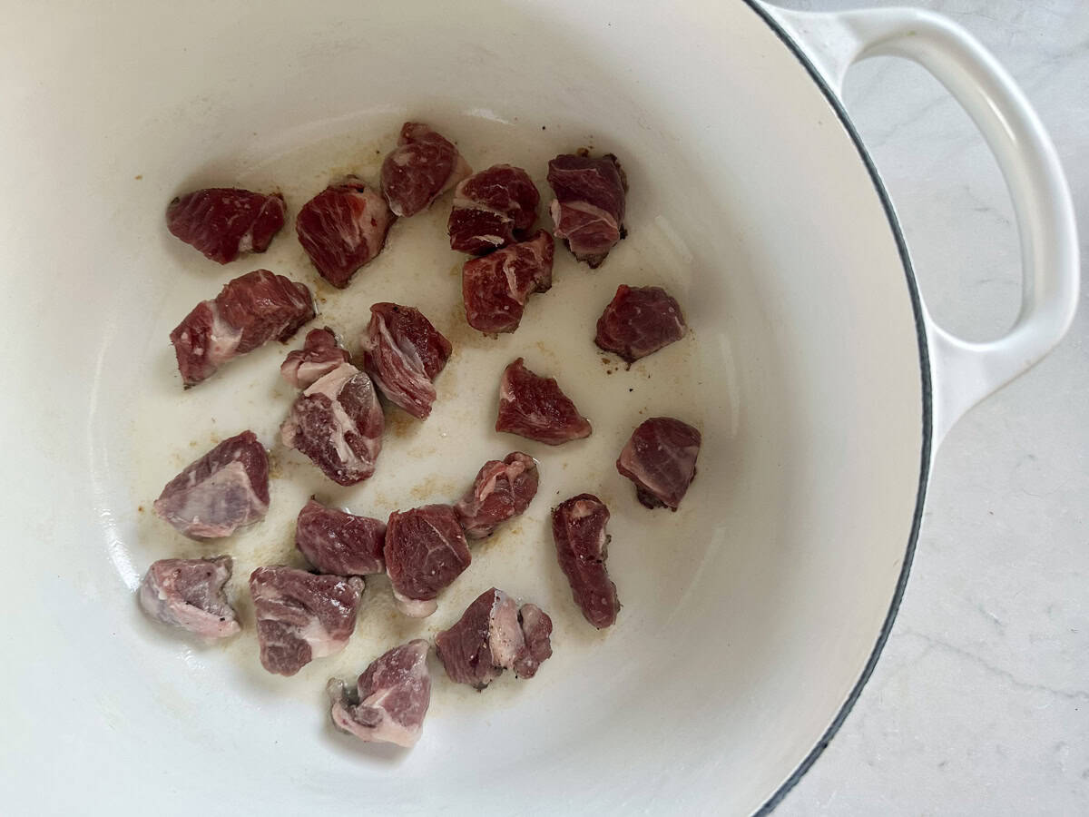 Chunks of raw beef are spread across the bottom of a white enameled pot, ready to be browned, on a light-colored countertop.